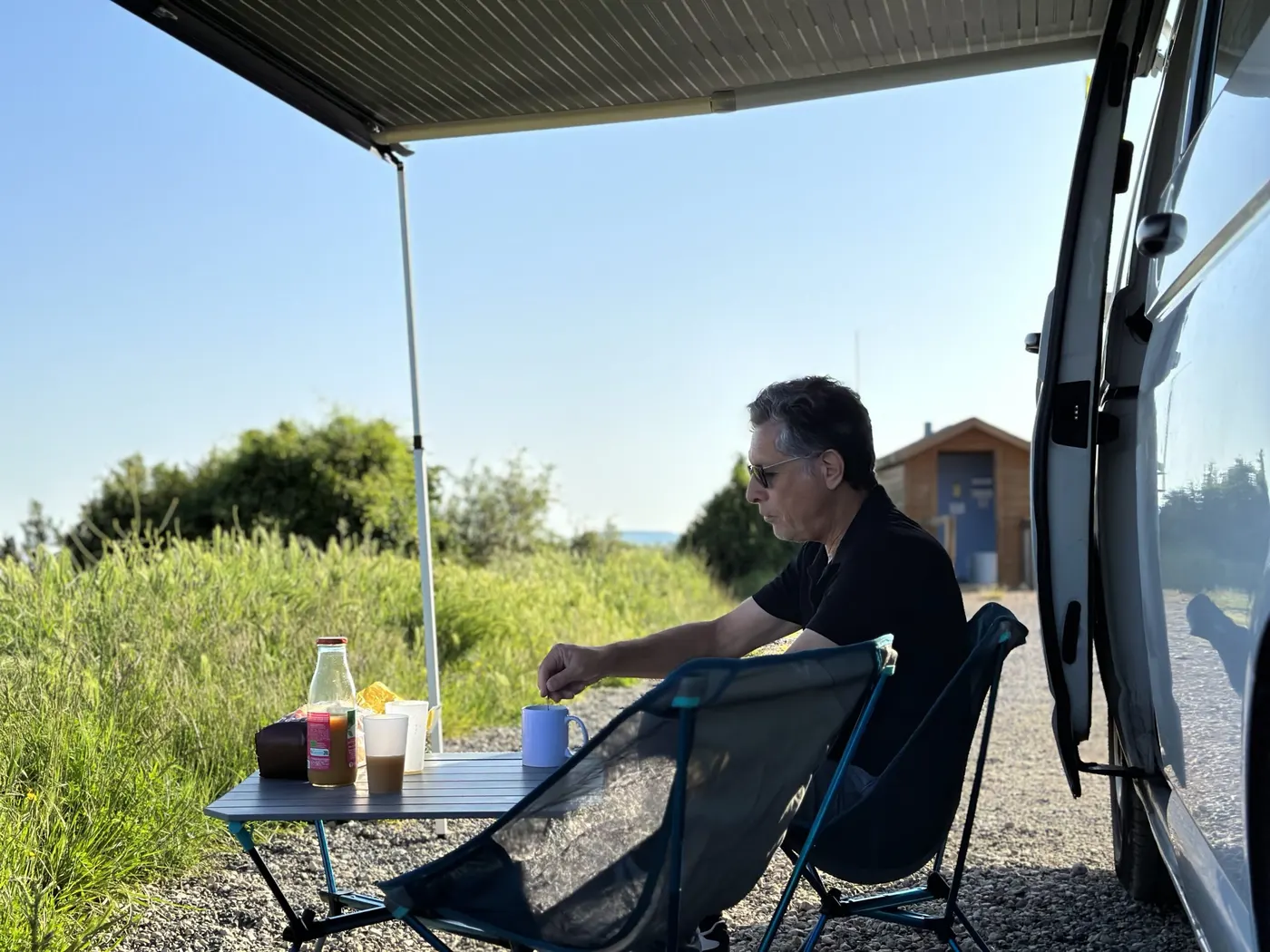 Un homme prend son café sous l’auvent de Jane, assis à une table de camping