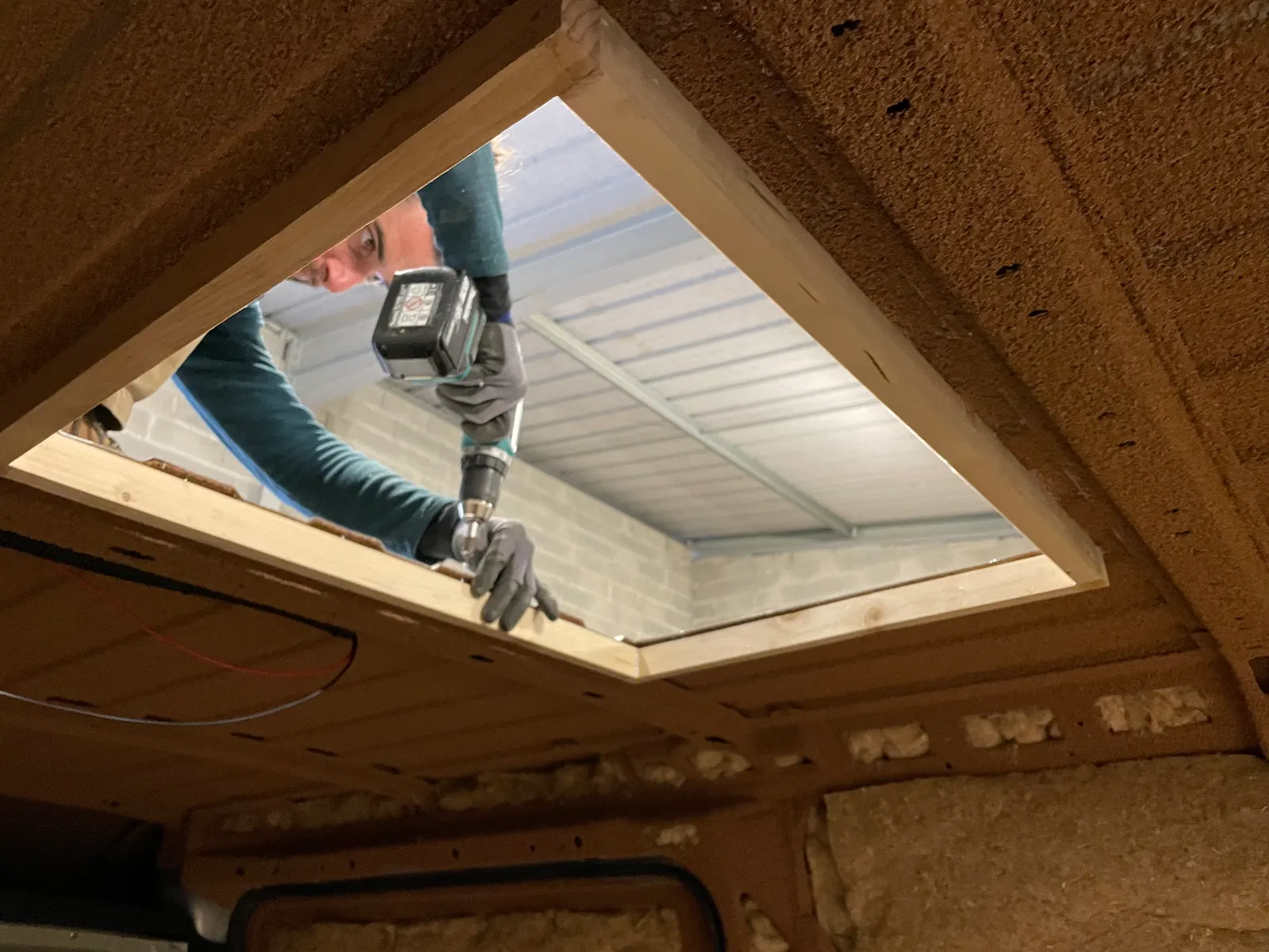 Vu de l'intérieur, un homme visse le cadre en bois d'un lanterneau au plafond d'un fourgon isolé