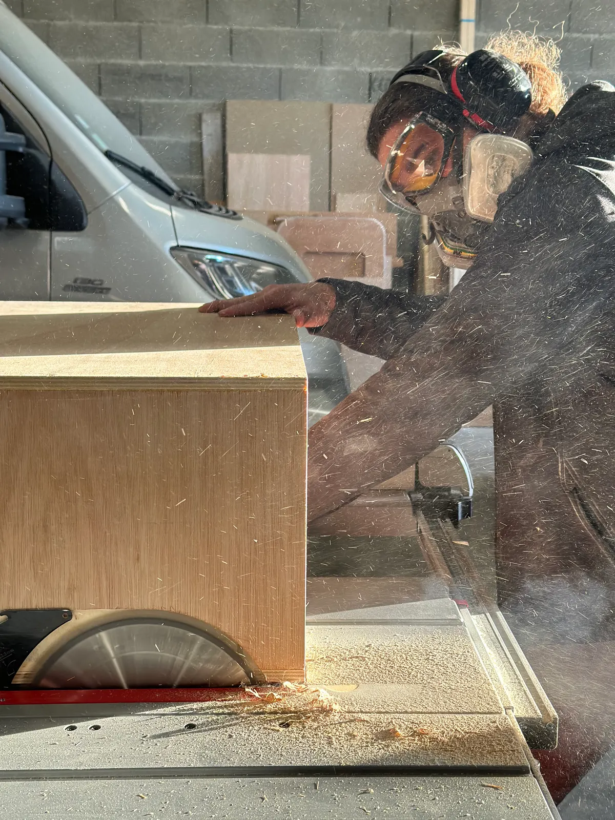 Un homme en masque et casque antibruit coupe un caisson en contreplaqué sur une scie sur table