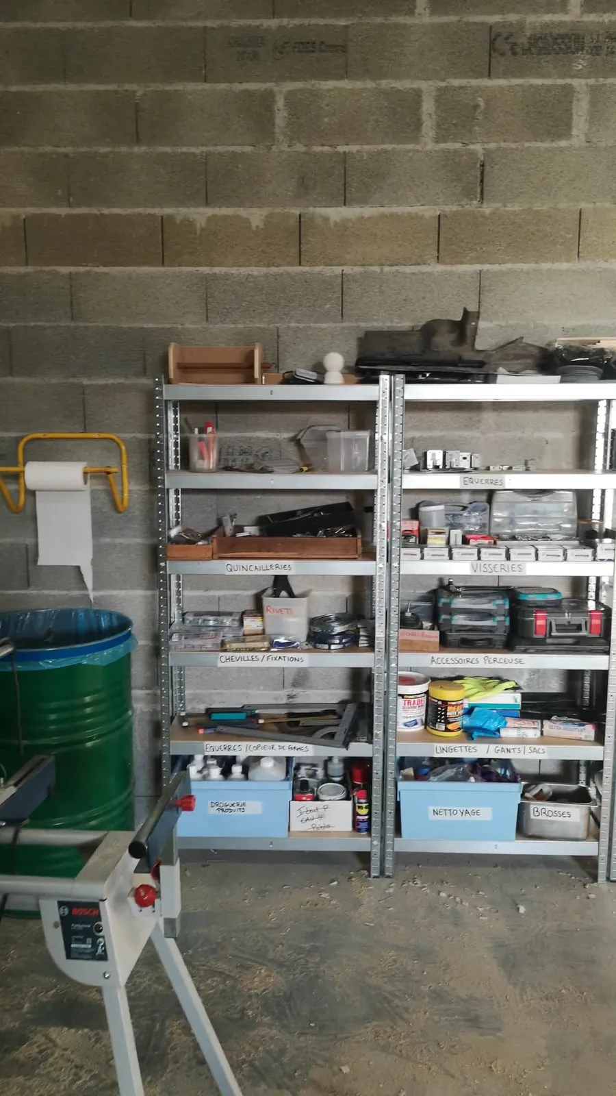 Étagères de l'atelier Le Van Trotter étiquetées par famille (quincailleries, visseries, chevilles et fixations, accessoires perceuse, équerres et copieur de formes, nettoyage), le piétement d'une scie Bosch au premier plan